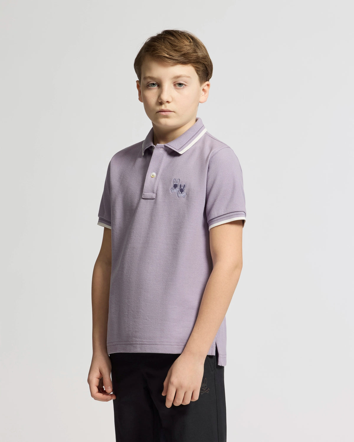 A young boy with short hair stands against a white background, wearing the stylish KIDS ELLIOT PIQUE POLO by Psycho Bunny in light purple. It's crafted from soft Pima cotton and features a small embroidered emblem on the chest. He gazes forward with a neutral expression.