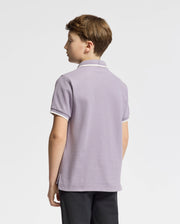 A young person with short brown hair stands facing away, wearing a stylish Psycho Bunny KIDS ELLIOT PIQUE POLO in lavender, made of soft Pima cotton, paired with black pants against a simple white background.