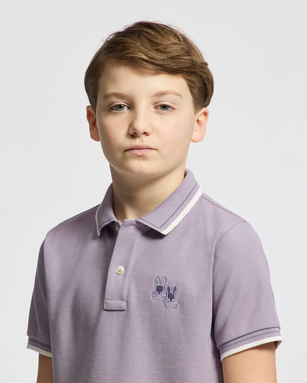 A young person with short light brown hair sports the stylish KIDS ELLIOT PIQUE POLO - B0K505F200 from Psycho Bunny, crafted from soft Pima cotton in light purple and featuring a small embroidered design. The background is plain white.