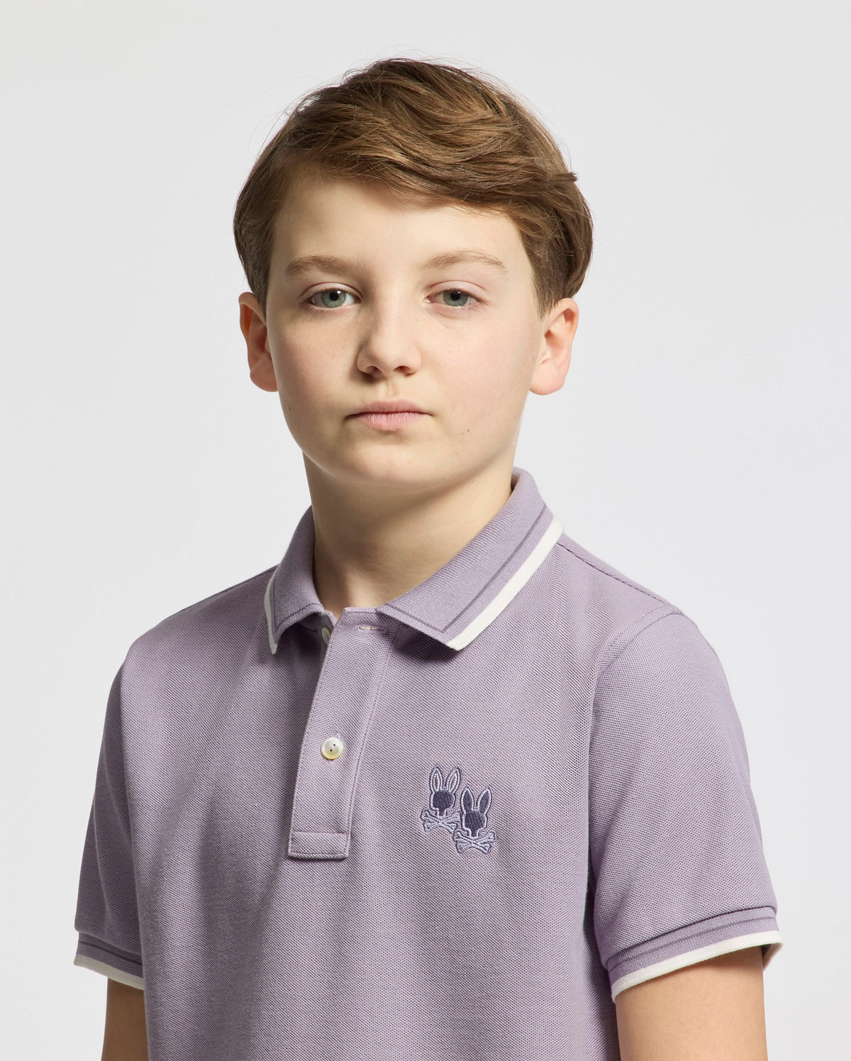 A young person with short light brown hair sports the stylish KIDS ELLIOT PIQUE POLO - B0K505F200 from Psycho Bunny, crafted from soft Pima cotton in light purple and featuring a small embroidered design. The background is plain white.
