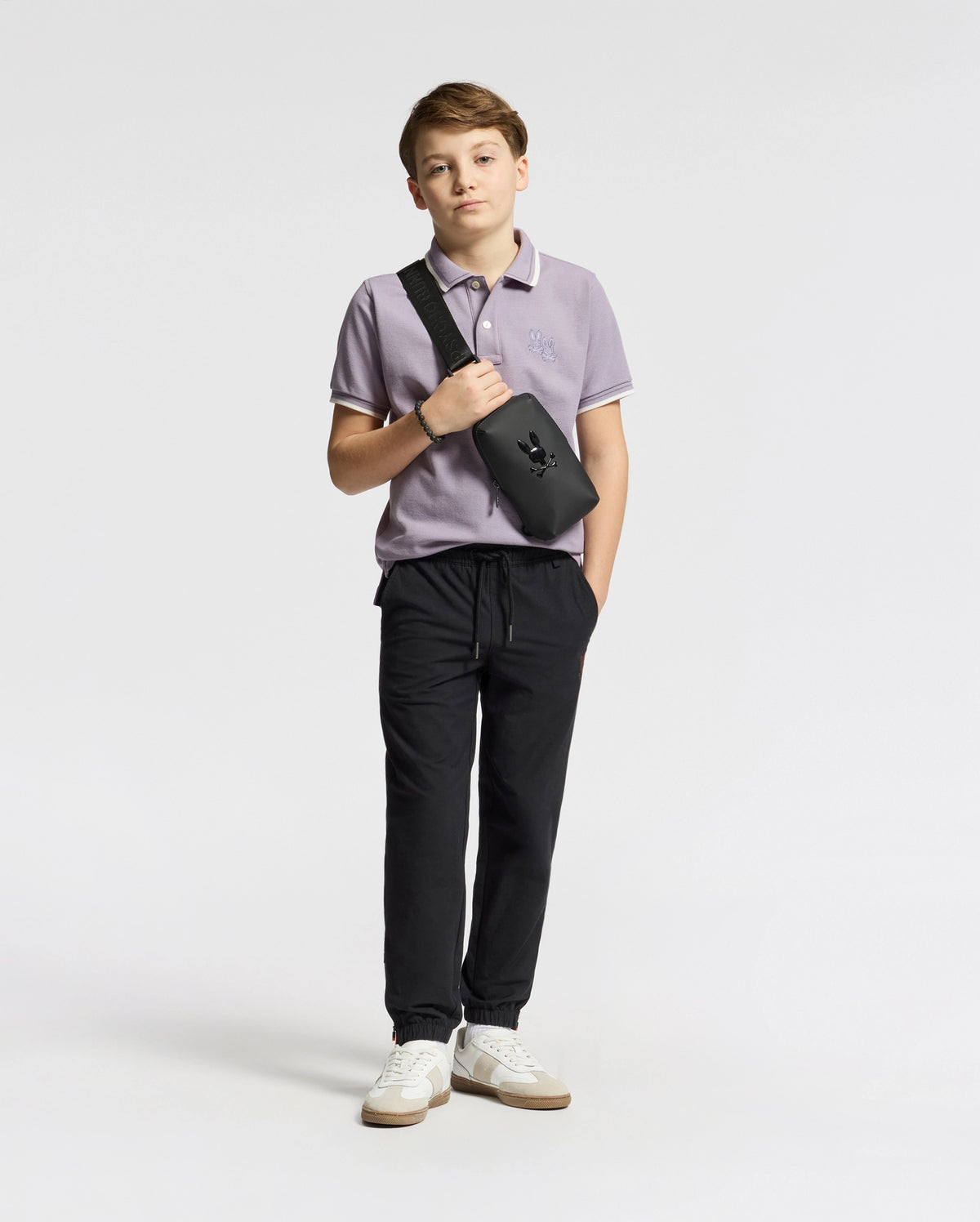 A trendy teen boy poses against a white backdrop in a lavender Psycho Bunny KIDS ELLIOT PIQUE POLO, black jogger pants, and white sneakers. He casually places his hands in his pockets while sporting a black crossbody bag over his shoulder.