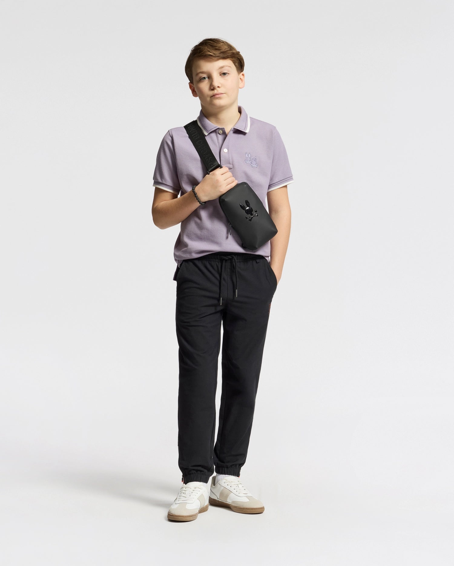 A trendy teen boy poses against a white backdrop in a lavender Psycho Bunny KIDS ELLIOT PIQUE POLO, black jogger pants, and white sneakers. He casually places his hands in his pockets while sporting a black crossbody bag over his shoulder.
