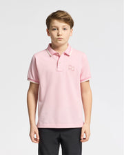 A young boy wearing the light pink KIDS ELLIOT PIQUE POLO by Psycho Bunny, made of soft Pima cotton and featuring a bold design with a small embroidered logo, stands against a white background. He has short brown hair, a neutral expression, and dark pants.