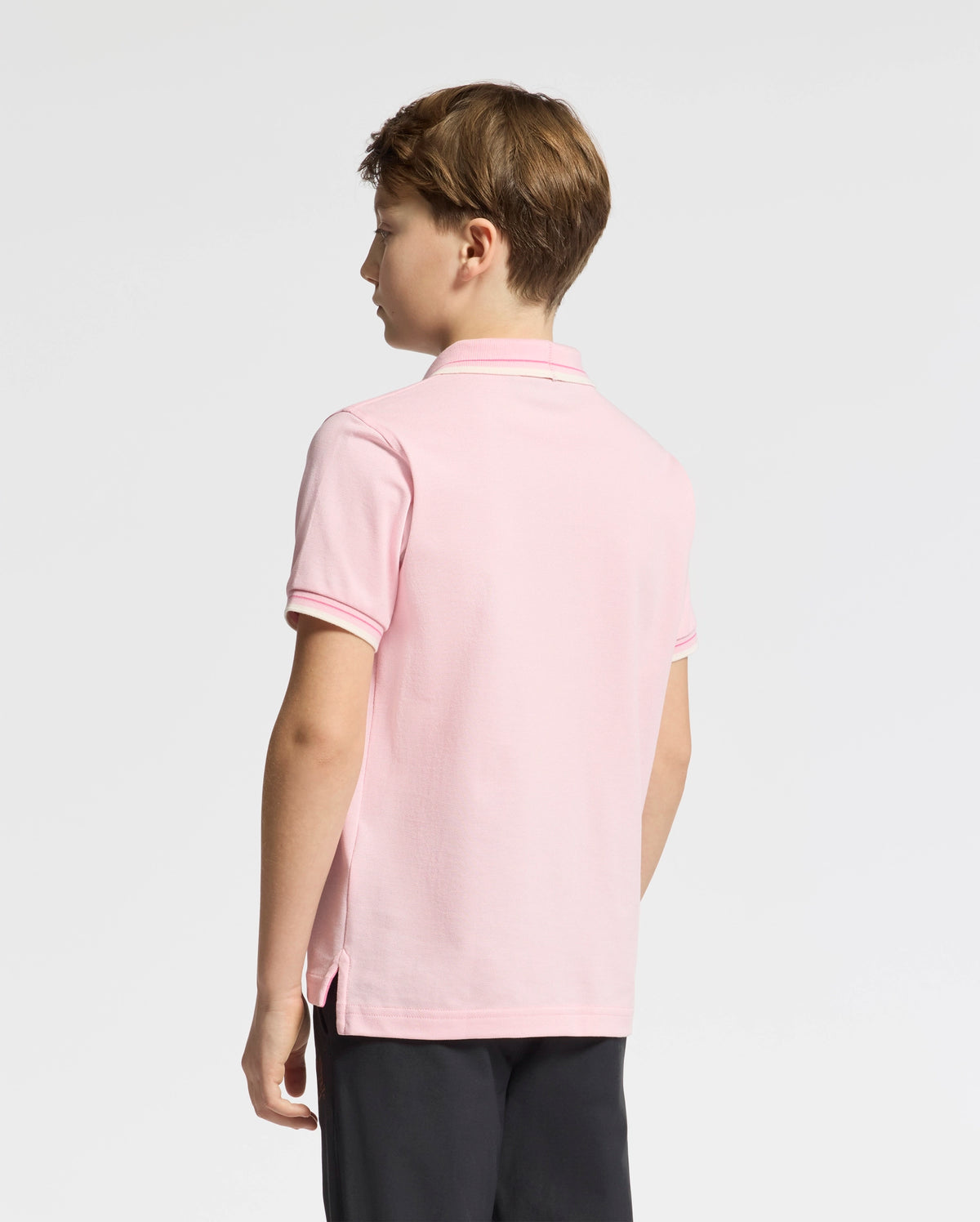A boy with brown hair stands sideways, facing left. He's wearing Psycho Bunny's Kids Elliot Pique Polo (B0K505F200) in light pink, made of soft Pima cotton, paired with black pants against a plain white background that highlights his bold outfit.