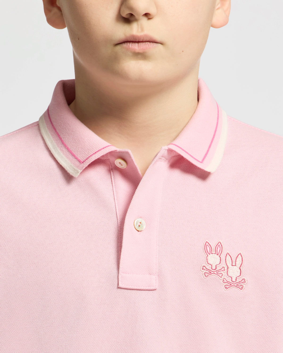 The child is wearing Psycho Bunny's Kids Elliot Pique Polo, a striking pink design with a collar featuring white and pink stripes. Made from soft Pima cotton, it includes two embroidered bunny logos on the chest for playful charm.