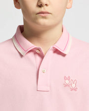 The child is wearing Psycho Bunny's Kids Elliot Pique Polo, a striking pink design with a collar featuring white and pink stripes. Made from soft Pima cotton, it includes two embroidered bunny logos on the chest for playful charm.