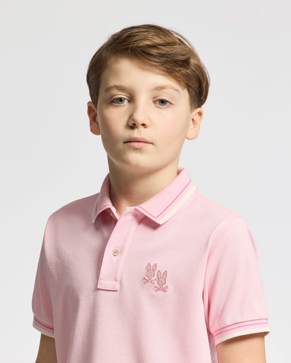 A boy with short brown hair wears a Psycho Bunny KIDS ELLIOT PIQUE POLO in pink, made from soft Pima cotton, featuring an embroidered logo on the chest. The simple white or light gray background highlights him.
