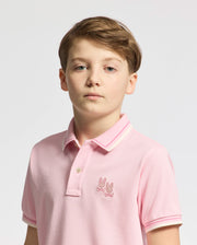 A boy with short brown hair wears a Psycho Bunny KIDS ELLIOT PIQUE POLO in pink, made from soft Pima cotton, featuring an embroidered logo on the chest. The simple white or light gray background highlights him.