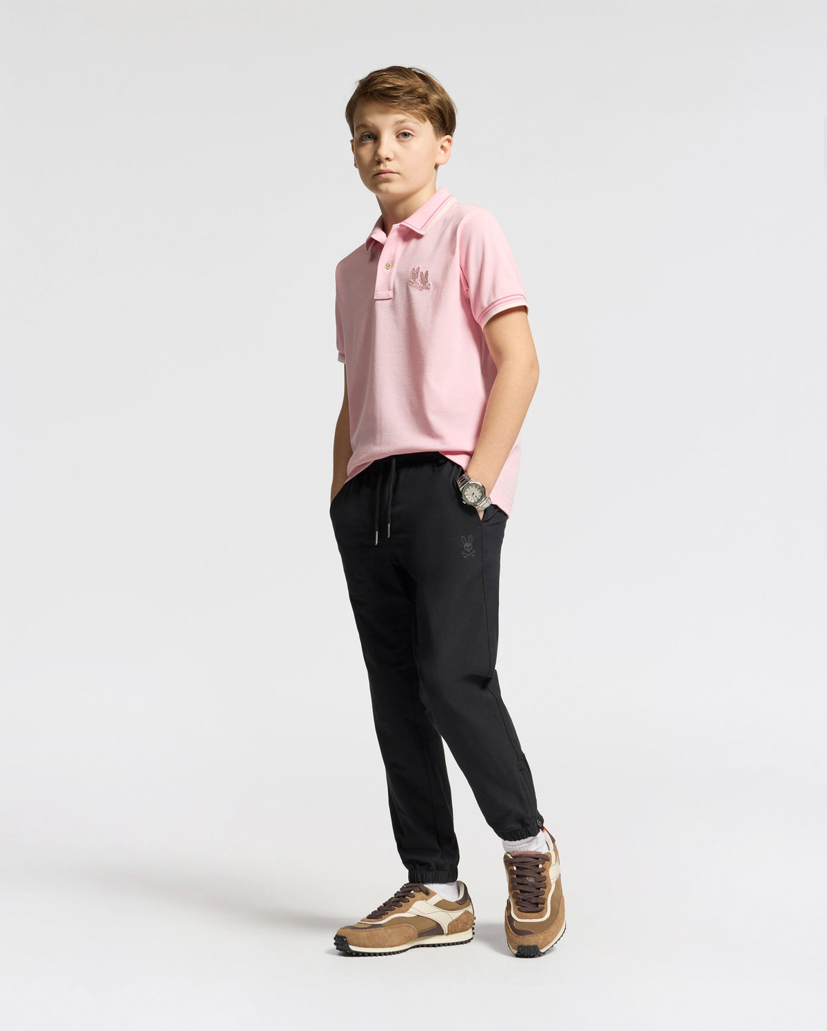 A boy confidently poses against a neutral background, wearing the Psycho Bunny KIDS ELLIOT PIQUE POLO - B0K505F200 in pink. He pairs it with black pants and brown sneakers, hands in pockets and boldly gazing at the camera.