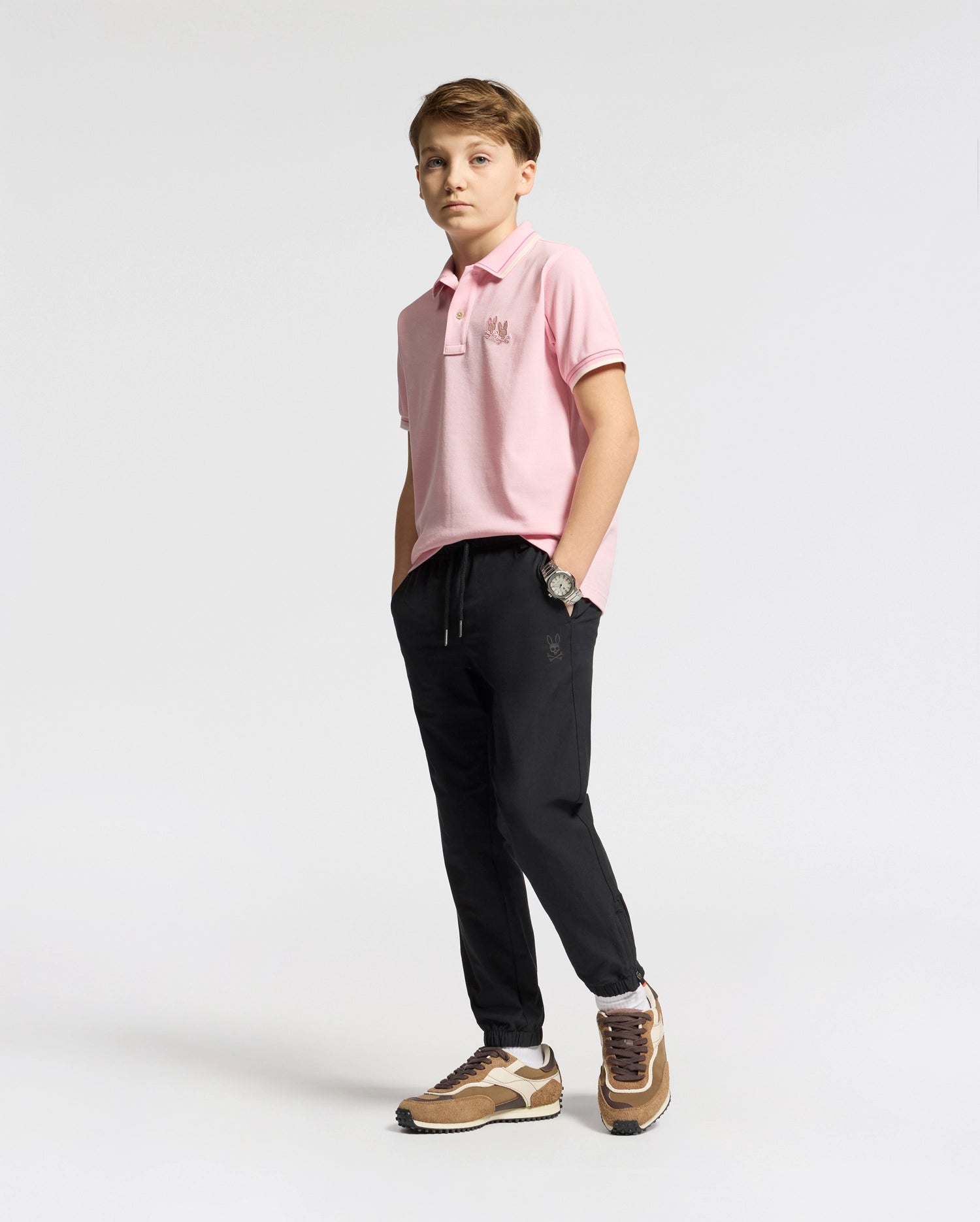 A boy confidently poses against a neutral background, wearing the Psycho Bunny KIDS ELLIOT PIQUE POLO - B0K505F200 in pink. He pairs it with black pants and brown sneakers, hands in pockets and boldly gazing at the camera.