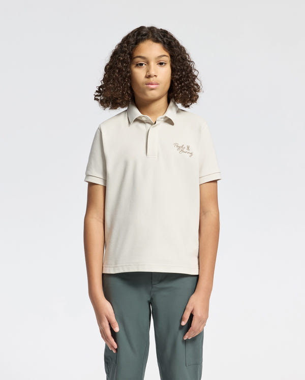A person with curly hair stands facing forward against a plain background, wearing a Psycho Bunny KIDS DESERT PIQUE POLO in soft Pima cotton. The shirt has chest text and pairs elegantly with dark green pants. Their arms rest relaxed by their sides, exuding timeless style.