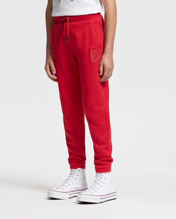 A person wearing Psycho Bunny's KIDS PIERCE ESSENTIAL FRENCH TERRY SWEATPANT - B0P136E200, featuring a small embroidered design on the side, and a white T-shirt stands against a plain background. They are also wearing white high-top sneakers with their hands resting by their sides.
