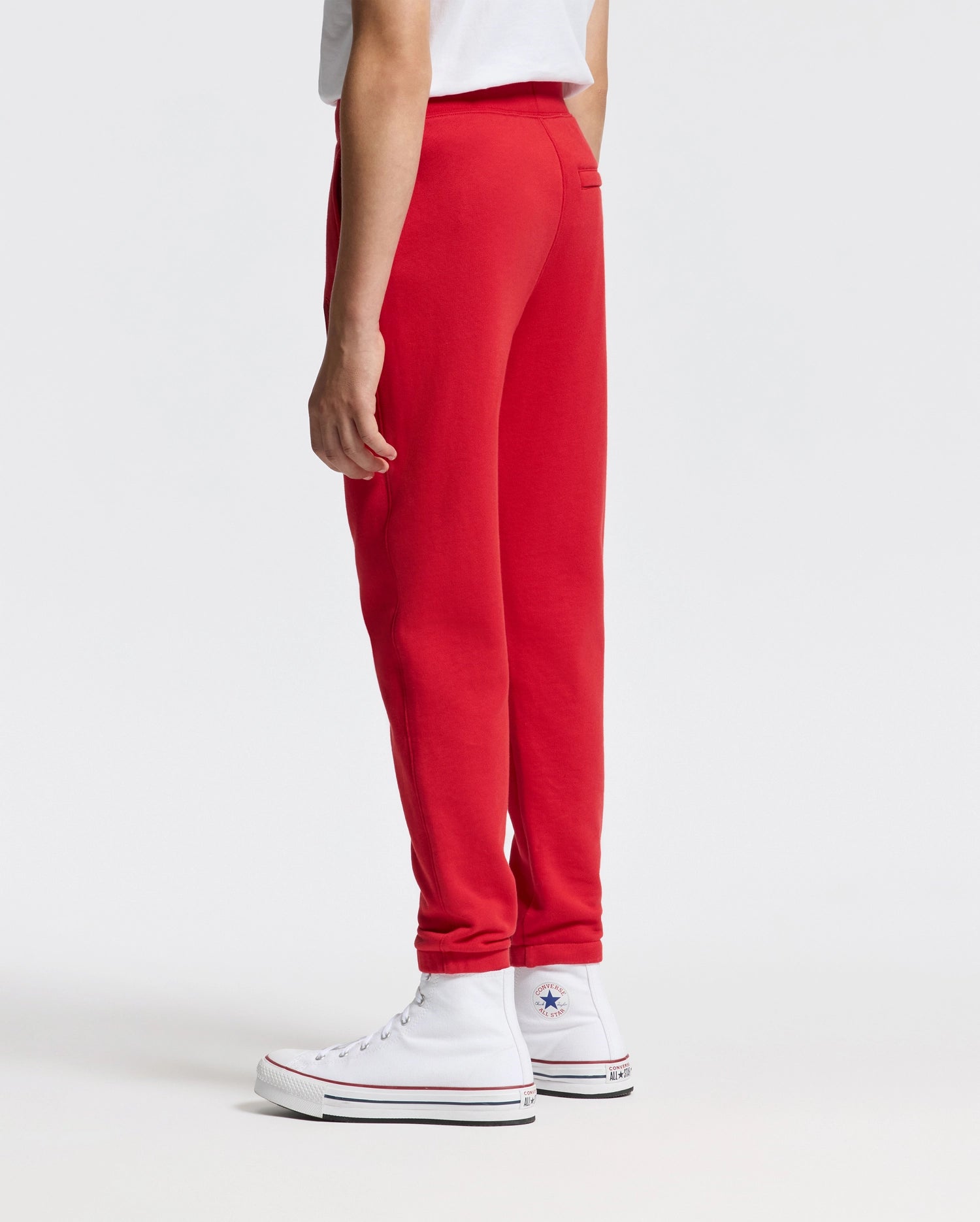 In a minimalist white setting, an individual showcases a striking ensemble featuring a white t-shirt and Psycho Bunny's vibrant red Kids Pierce Essential French Terry Sweatpants (B0P136E200). The look is complemented by high-top sneakers embellished with star logos.