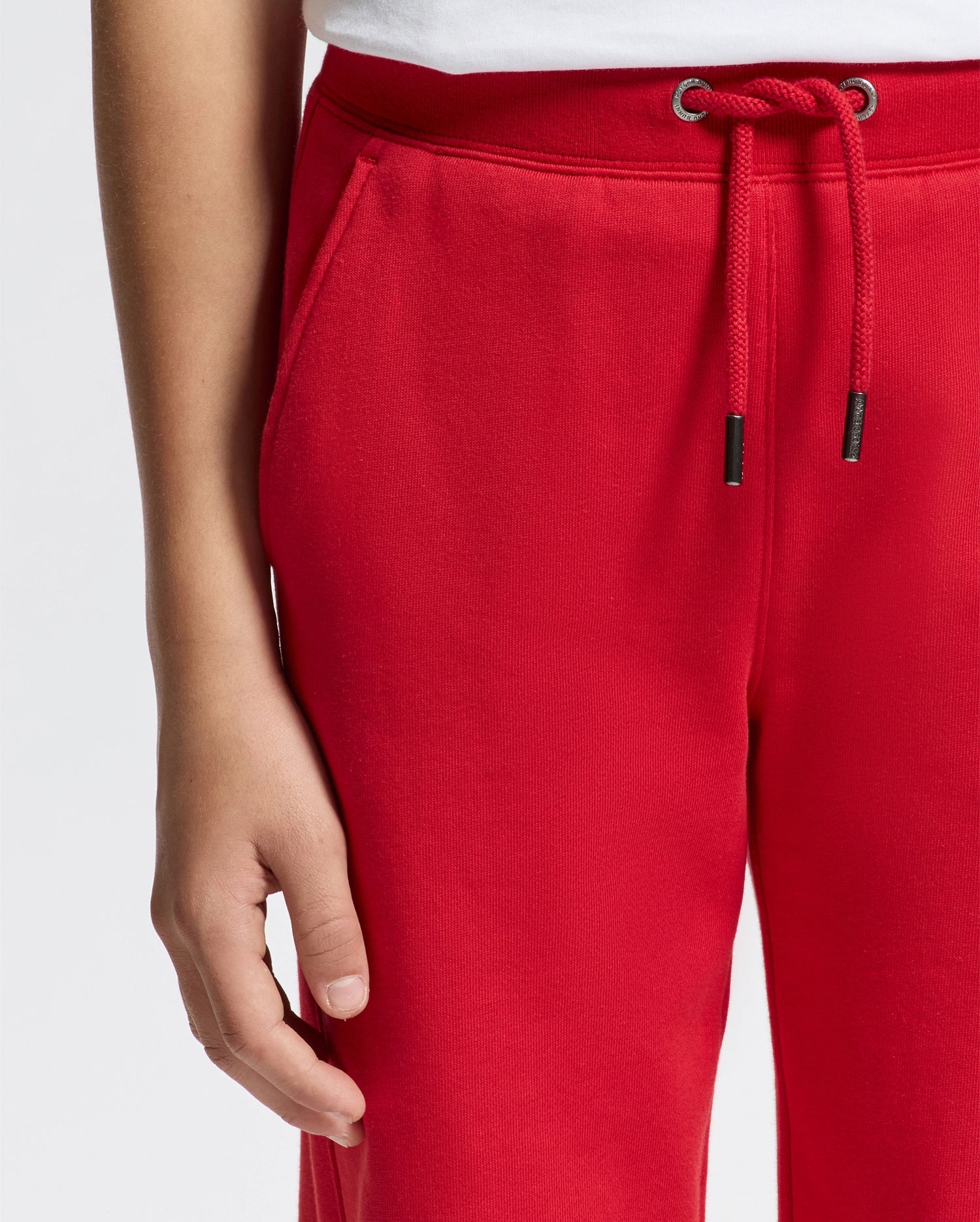 A person wearing bright red, premium quality KIDS PIERCE ESSENTIAL FRENCH TERRY SWEATPANT by Psycho Bunny with a drawstring is standing against a light background. The left hand is visible, resting by their side. The sweatpants feature side pockets, and the hem of a white shirt is slightly visible.