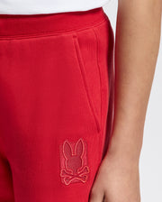 A person wearing Psycho Bunny's KIDS PIERCE ESSENTIAL FRENCH TERRY SWEATPANT in red, featuring a stitched rabbit and crossbones logo. These premium quality kids' joggers include a visible side pocket for added convenience.