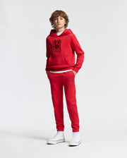 A young person wearing a red hoodie and matching Psycho Bunny KIDS PIERCE ESSENTIAL FRENCH TERRY SWEATPANT - B0P136E200 stands against a plain white background. The hoodie displays a graphic on the front, paired perfectly with their French terry sweatpants and white high-top sneakers.