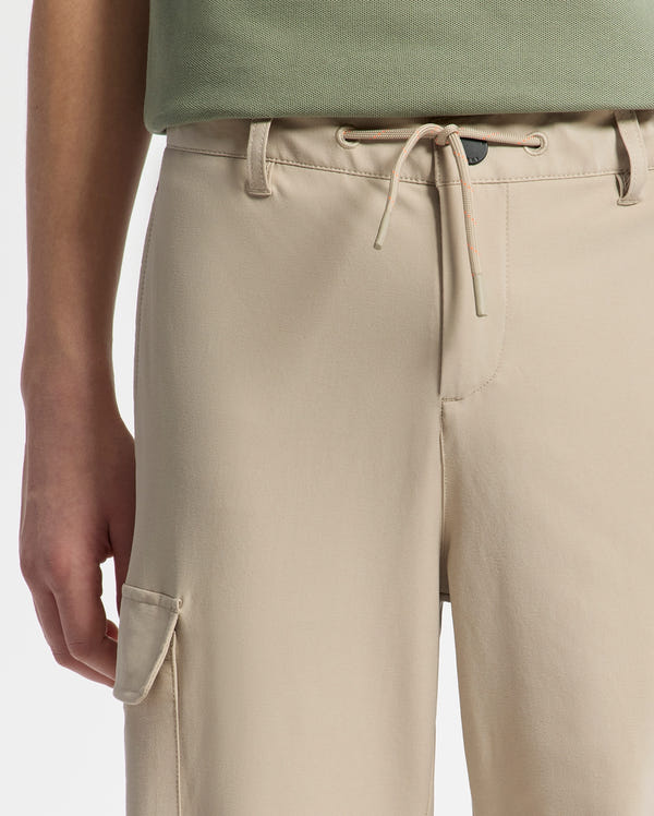 A person is wearing beige Psycho Bunny KIDS GREEN VALLEY CARGO PANT - B0P199E200 with an elastic waistband, drawstring, and side pocket. They also have a green top that is partly visible as the focus is on the waist and upper thigh of the pants.