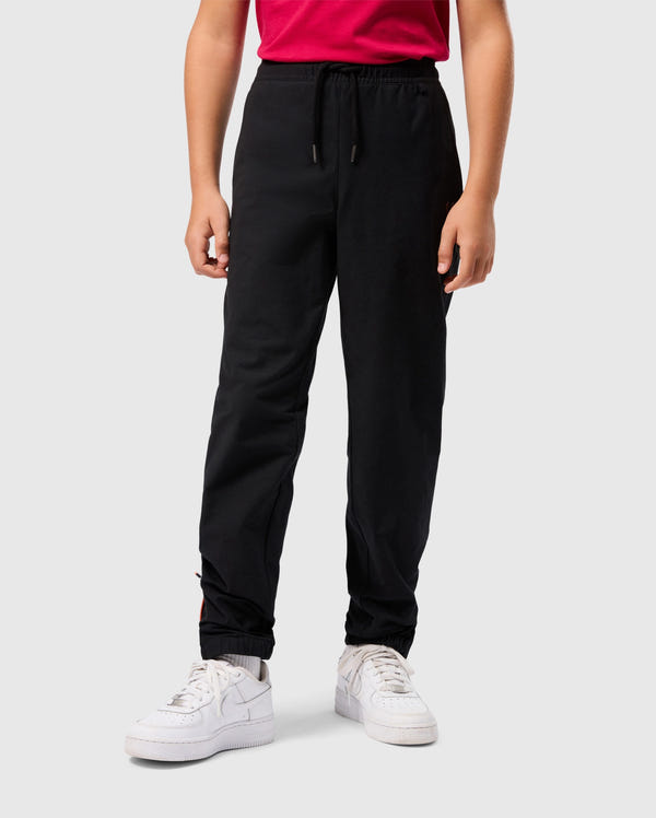 A person wearing a red shirt, KIDS MADISON COMMUTER PANT - B0P395Z1WB with zippered expansion vents, and white sneakers is standing against a plain white background. Only the lower half of the body is visible in the image. The pant by Psycho Bunny features an elastic waistband and cuffs, adorned with a detachable Bunny keychain.