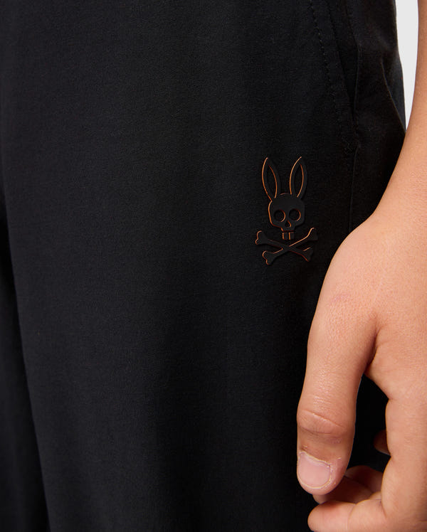 Close-up of a person's hand resting by the side of black Kids Madison Commuter Pant - B0P395Z1WB from Psycho Bunny. The pants feature a small emblem of a cartoon skull and crossbones with bunny ears on the thigh. Only part of the hand and pants are visible in the image, hinting at zippered expansion vents for added comfort.