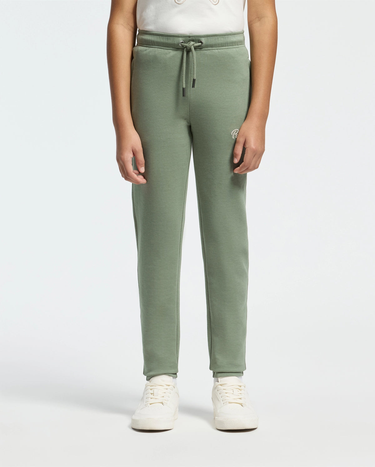 A person wearing the KIDS DESERT EMBROIDERED SWEATPANT by Psycho Bunny, crafted from a soft stretch jersey, alongside a white shirt and sneakers, stands against a plain light background.