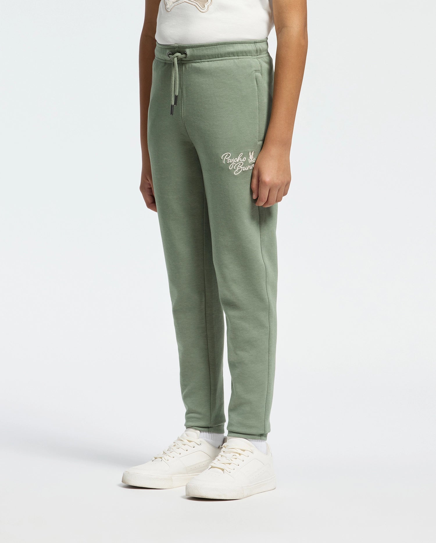 A person in Psycho Bunny's KIDS DESERT EMBROIDERED SWEATPANT (B0P489F200) in soft stretch jersey, featuring a drawstring waist and thigh logo, pairs them with white sneakers and a matching top against a plain background.