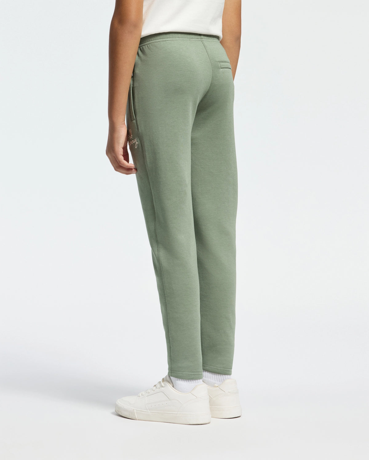 A person in a white shirt and sneakers stands sideways, wearing Psycho Bunny's Kids Desert Embroidered Sweatpant - B0P489F200 made from soft stretch jersey against a plain white background.
