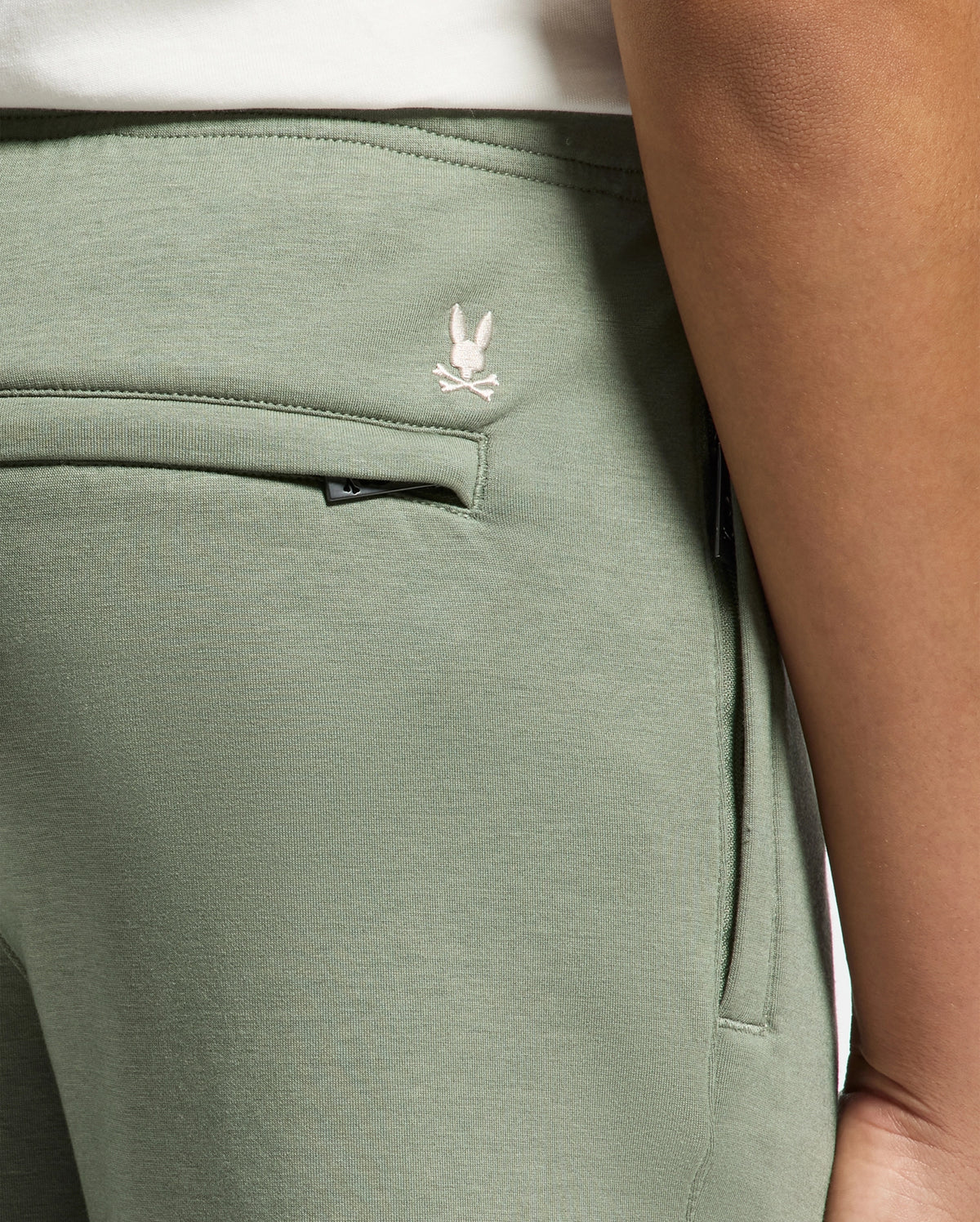 Close-up of a person wearing light green Psycho Bunny KIDS DESERT EMBROIDERED SWEATPANT - B0P489F200 made from soft stretch jersey, featuring a small white embroidered rabbit and crossed bones logo near the waistband. The image highlights the side pocket, seam detail, and Modal-blend comfort.