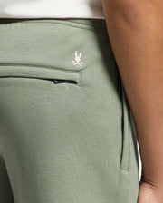 Close-up of a person wearing light green Psycho Bunny KIDS DESERT EMBROIDERED SWEATPANT - B0P489F200 made from soft stretch jersey, featuring a small white embroidered rabbit and crossed bones logo near the waistband. The image highlights the side pocket, seam detail, and Modal-blend comfort.