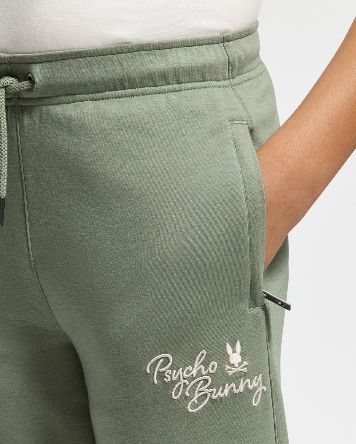 Close-up of someone wearing KIDS DESERT EMBROIDERED SWEATPANT in light green, made from soft stretch jersey with "Psycho Bunny" and a bunny logo embroidered on the pocket. A hand is partially tucked into the pocket, and a drawstring is visible on the left.
