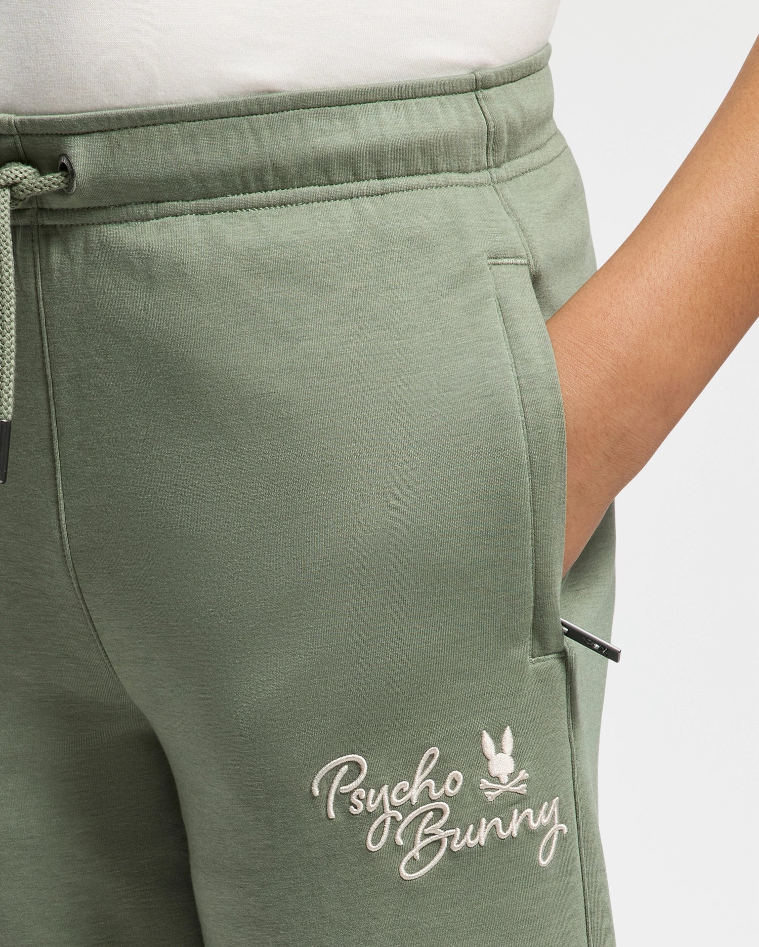Close-up of someone wearing KIDS DESERT EMBROIDERED SWEATPANT in light green, made from soft stretch jersey with "Psycho Bunny" and a bunny logo embroidered on the pocket. A hand is partially tucked into the pocket, and a drawstring is visible on the left.