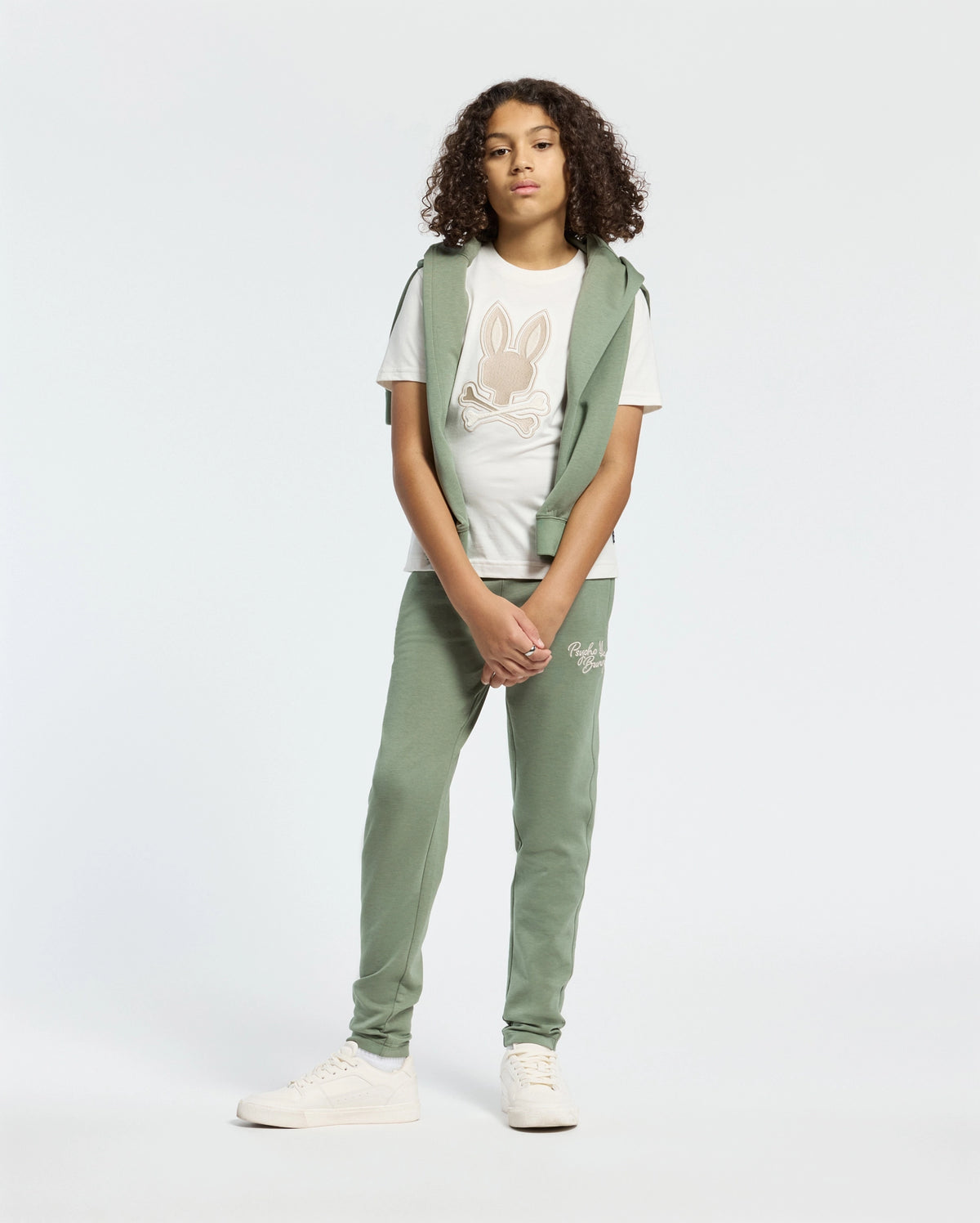 A person with curly hair wears a light green hoodie, a white T-shirt with a rabbit design, and matching KIDS DESERT EMBROIDERED SWEATPANTS by Psycho Bunny. They finish the look with white sneakers against a plain white background.