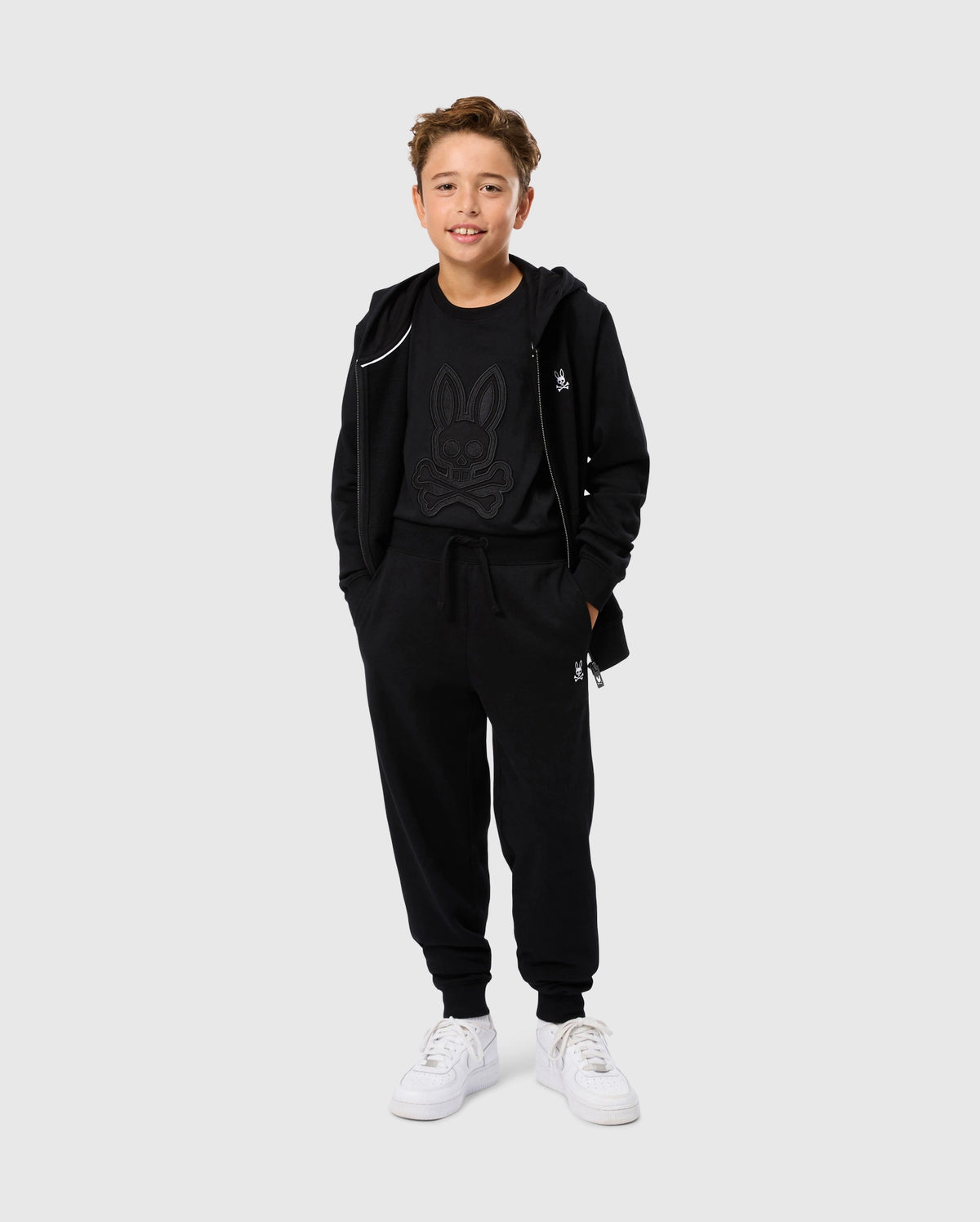 A young person with short hair smiles while wearing a black hoodie, a graphic t-shirt featuring a stylized bunny, and matching KIDS LEVY ESSENTIAL FLEECE SWEATPANTS (B0P721D200) from Psycho Bunny. They are also wearing white sneakers. The background is plain light gray.
