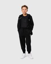 A young person with short hair smiles while wearing a black hoodie, a graphic t-shirt featuring a stylized bunny, and matching KIDS LEVY ESSENTIAL FLEECE SWEATPANTS (B0P721D200) from Psycho Bunny. They are also wearing white sneakers. The background is plain light gray.