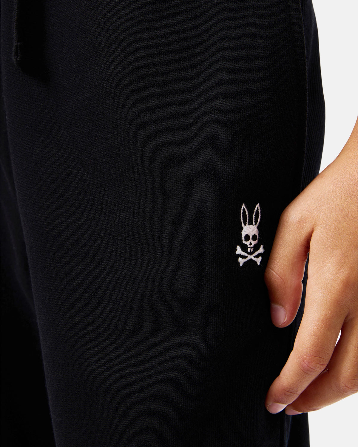 Close-up of a person's hand resting on their leg, wearing the KIDS LEVY ESSENTIAL FLEECE SWEATPANT - B0P721D200 by Psycho Bunny. The black fabric showcases an embroidered white logo of a rabbit head with crossbones near the pocket. These 100% cotton pants offer both comfort and style.