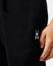 Close-up of a person's hand resting on their leg, wearing the KIDS LEVY ESSENTIAL FLEECE SWEATPANT - B0P721D200 by Psycho Bunny. The black fabric showcases an embroidered white logo of a rabbit head with crossbones near the pocket. These 100% cotton pants offer both comfort and style.