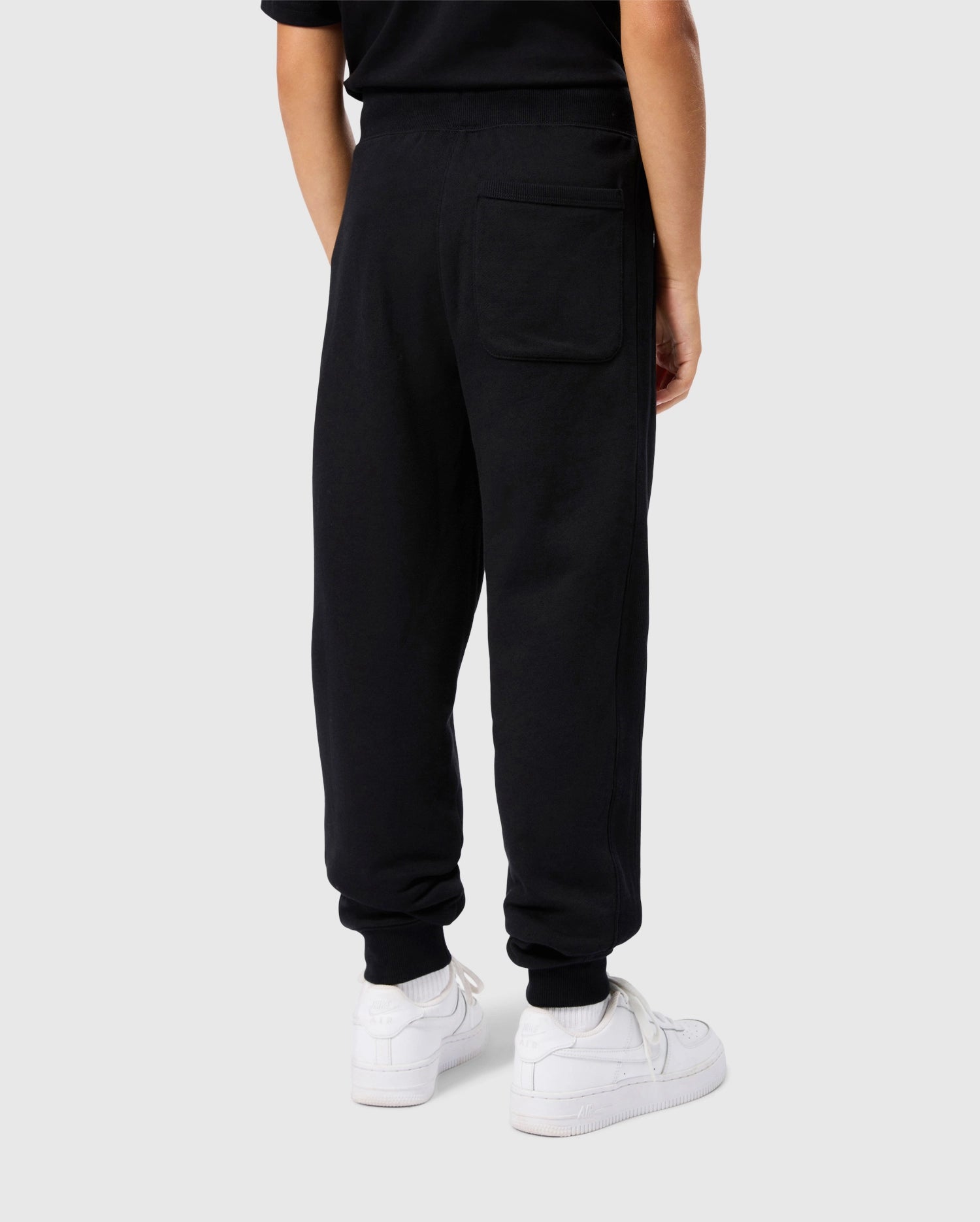 A person is seen from the back wearing a black T-shirt and Psycho Bunny's KIDS LEVY ESSENTIAL FLEECE SWEATPANT - B0P721D200, featuring a large pocket on the right side. The cozy playtime pants complement their white sneakers perfectly, all against a plain gray background.