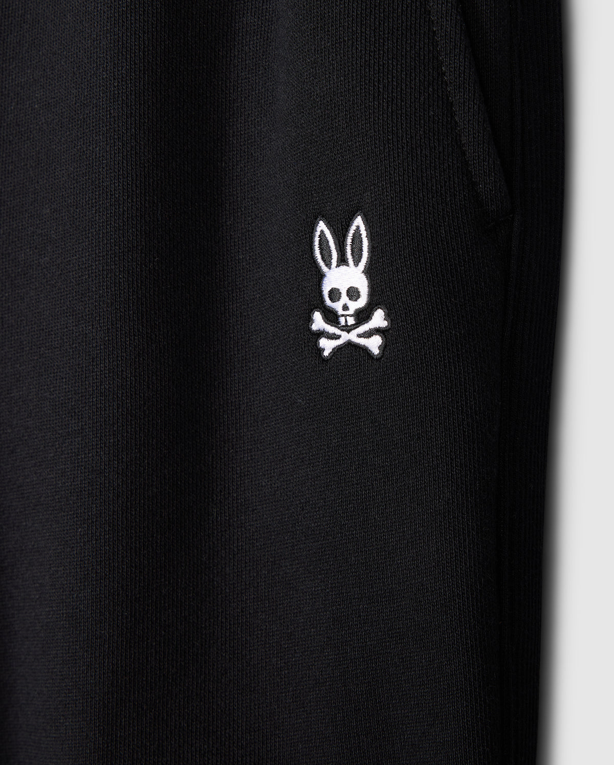 Close-up of a black fabric featuring a distinctive white embroidered logo from Psycho Bunny. The logo showcases a cartoonish bunny head with long ears above a skull and crossbones design, blending playful and edgy elements. This fabric appears to be part of the KIDS LEVY ESSENTIAL FLEECE SWEATPANT - B0P721D200, cozy playtime pants for kids.