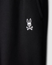 Close-up of a black fabric featuring a distinctive white embroidered logo from Psycho Bunny. The logo showcases a cartoonish bunny head with long ears above a skull and crossbones design, blending playful and edgy elements. This fabric appears to be part of the KIDS LEVY ESSENTIAL FLEECE SWEATPANT - B0P721D200, cozy playtime pants for kids.