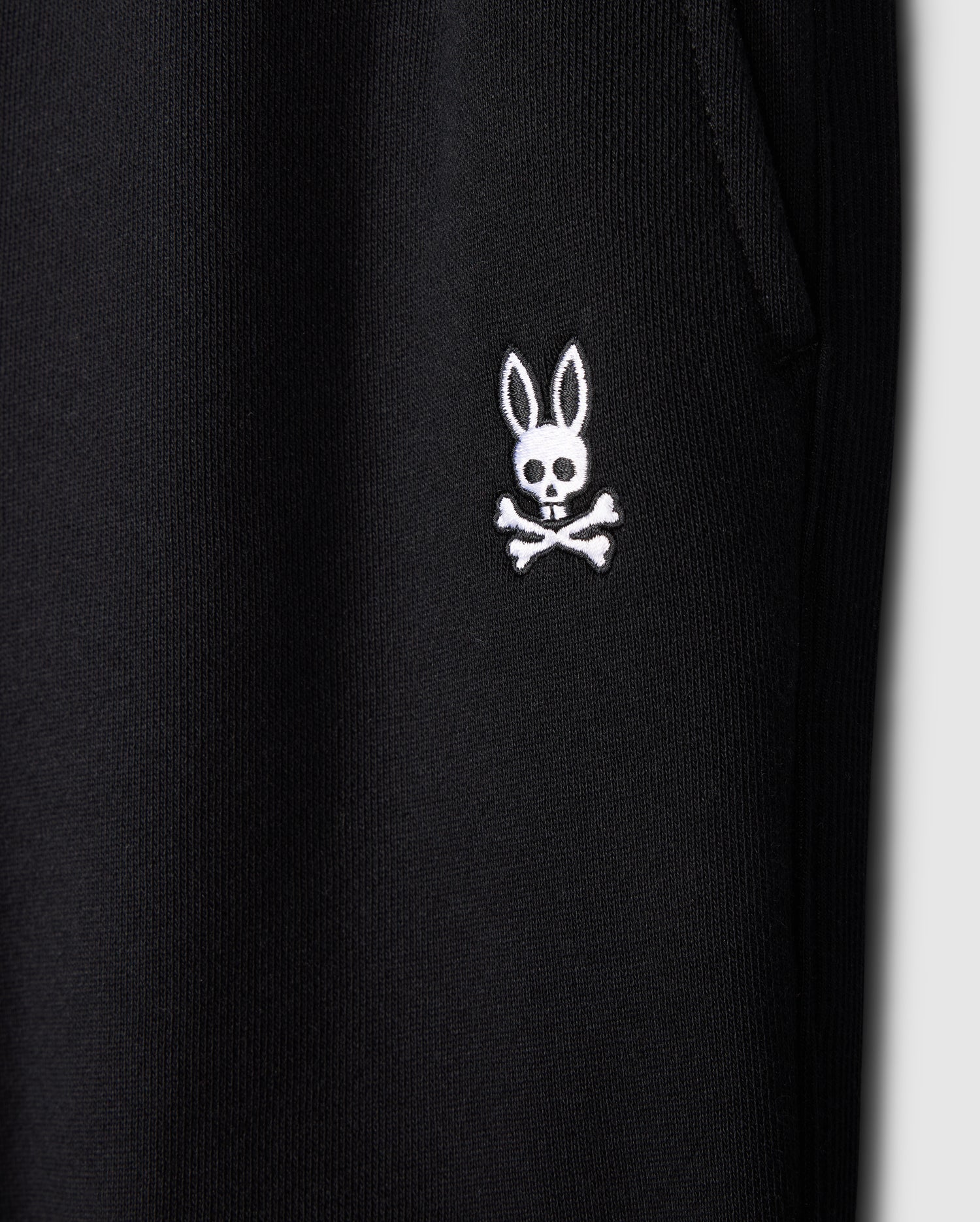 Close-up of a black fabric featuring a distinctive white embroidered logo from Psycho Bunny. The logo showcases a cartoonish bunny head with long ears above a skull and crossbones design, blending playful and edgy elements. This fabric appears to be part of the KIDS LEVY ESSENTIAL FLEECE SWEATPANT - B0P721D200, cozy playtime pants for kids.