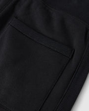Close-up of the KIDS LEVY ESSENTIAL FLEECE SWEATPANT - B0P721D200 by Psycho Bunny in black fabric, featuring a patch pocket. The texture of the 100% cotton fabric and stitching detail on the pocket are clearly visible, with ribbed elements along the edges of the pocket. This design is perfect for those cozy playtime pants.