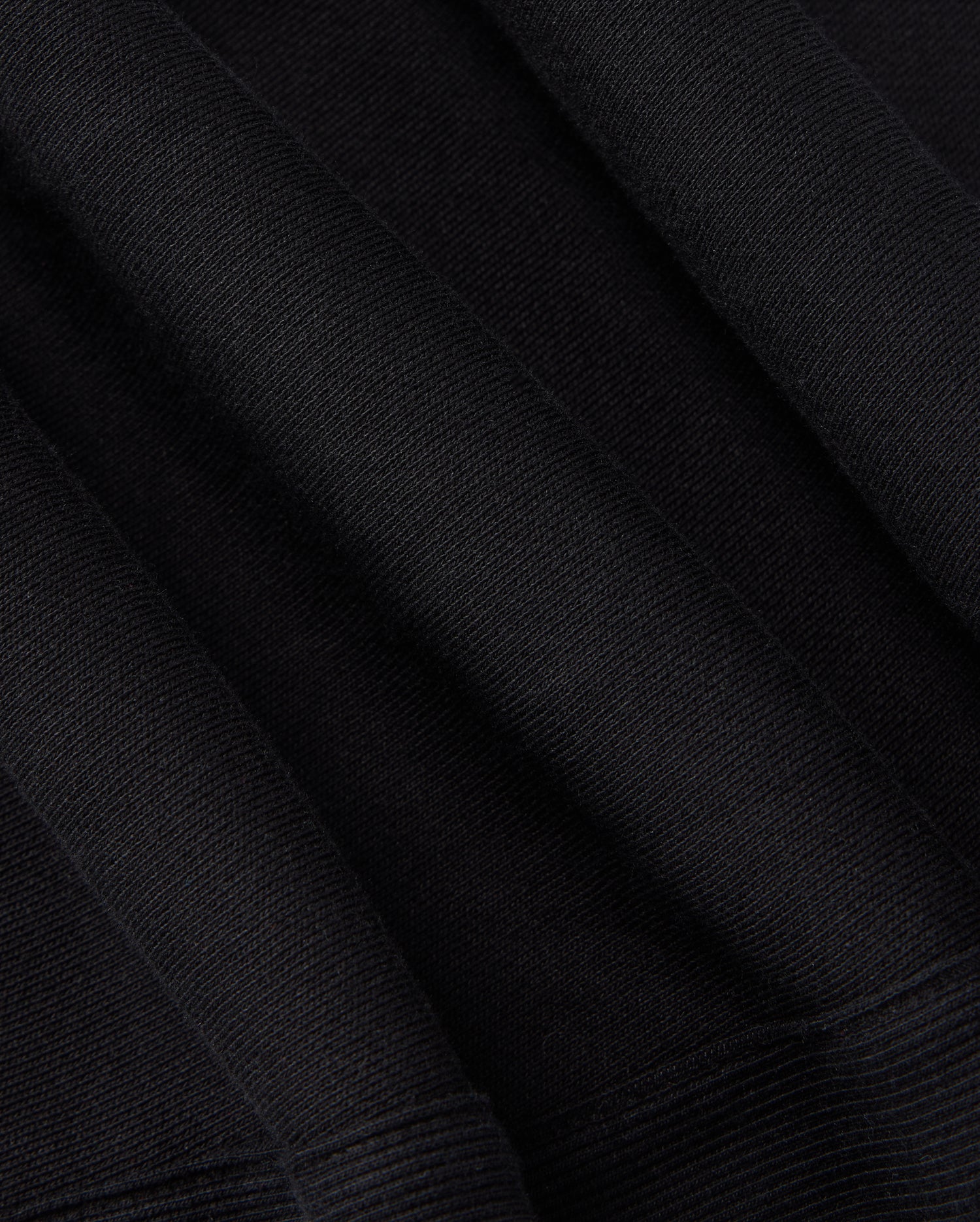 Close-up of the KIDS LEVY ESSENTIAL FLEECE SWEATPANT - B0P721D200 by Psycho Bunny. The black fabric has a ribbed texture and appears to have folds and creases, showcasing its thickness and material composition, much like cozy playtime pants. The texture suggests a soft and potentially warm fabric, ideal for clothing or upholstery needs.