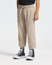 A person is sporting the KIDS LEVY ESSENTIAL FLEECE SWEATPANT - B0P721D200 in a cozy beige color, featuring a small Psycho Bunny logo on the left pocket. These are paired with black and white high-top sneakers, as they stand against a plain light background, radiating casual charm.