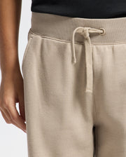 A person is depicted from the waist down wearing Psycho Bunny's KIDS LEVY ESSENTIAL FLEECE SWEATPANT in beige, featuring a drawstring and pockets. They are also paired with a black shirt, embodying a comfortable, casual vibe thanks to the soft fabric.