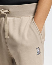 A close-up of a person wearing the KIDS LEVY ESSENTIAL FLEECE SWEATPANT - B0P721D200 by Psycho Bunny, showcasing its cozy beige design with one hand in the pocket. These cotton joggers come with a drawstring waistband and feature a small embroidered logo of a skull and crossbones with rabbit ears on the left side.