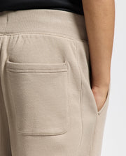 A close-up of a person wearing the KIDS LEVY ESSENTIAL FLEECE SWEATPANT in light beige from Psycho Bunny. The sweatpants are designed with a patch pocket on the back, while the person's right arm rests at their side. Crafted from soft, comfortable fabric, these cotton joggers blend style and ease for any casual occasion.