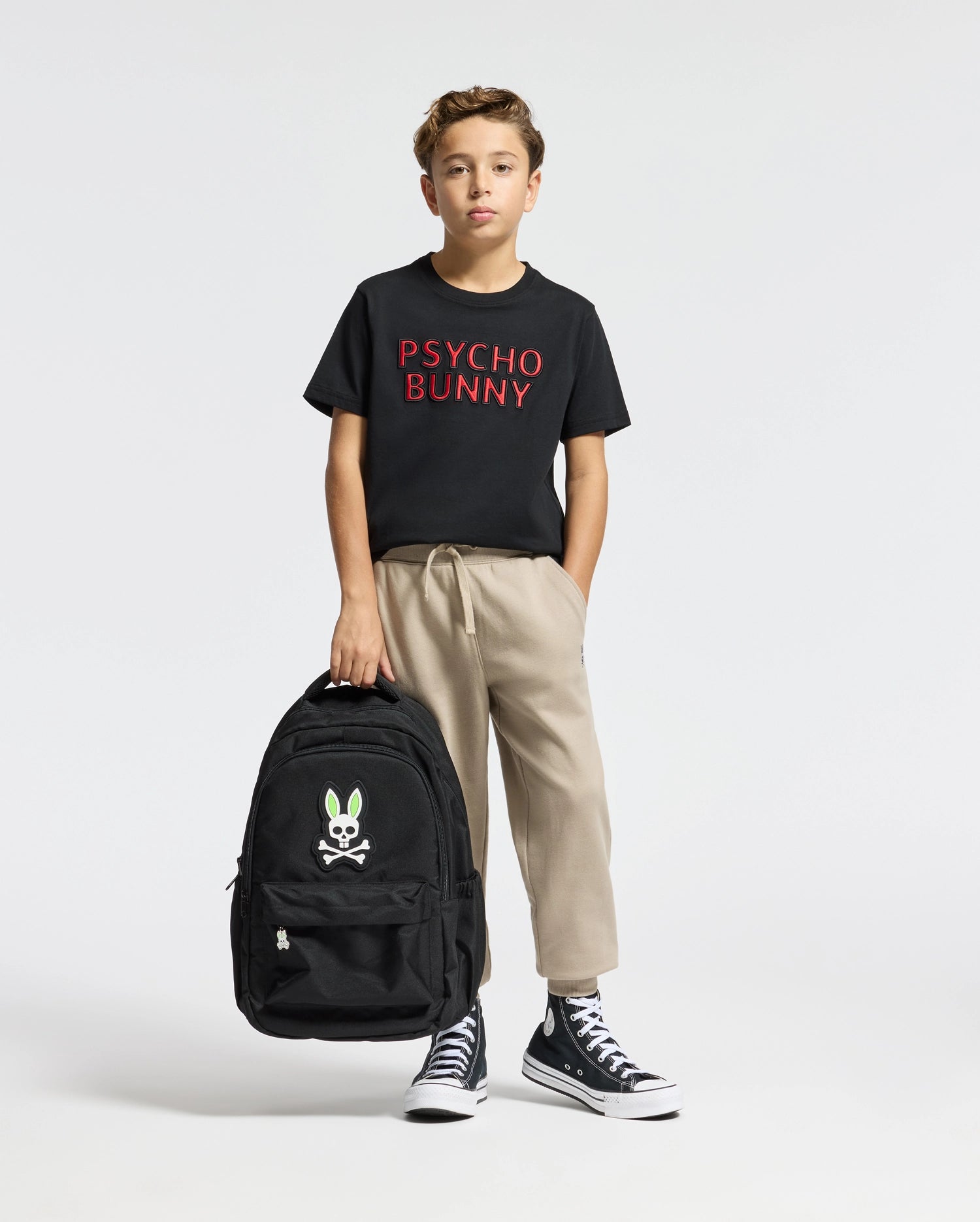 A young boy stands holding a large black backpack featuring a bunny and crossbones logo. He is dressed in a stylish Psycho Bunny black t-shirt with red lettering, cozy KIDS LEVY ESSENTIAL FLEECE SWEATPANT - B0P721D200, and black high-top sneakers.