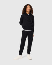 A person with curly hair is standing confidently against a plain background, wearing a black hoodie and the KIDS PACIFIC CHENILLE JOGGER - B0P972D200 by Psycho Bunny. They have their hands behind their back and are smiling, sporting casual bottoms paired with white sneakers.