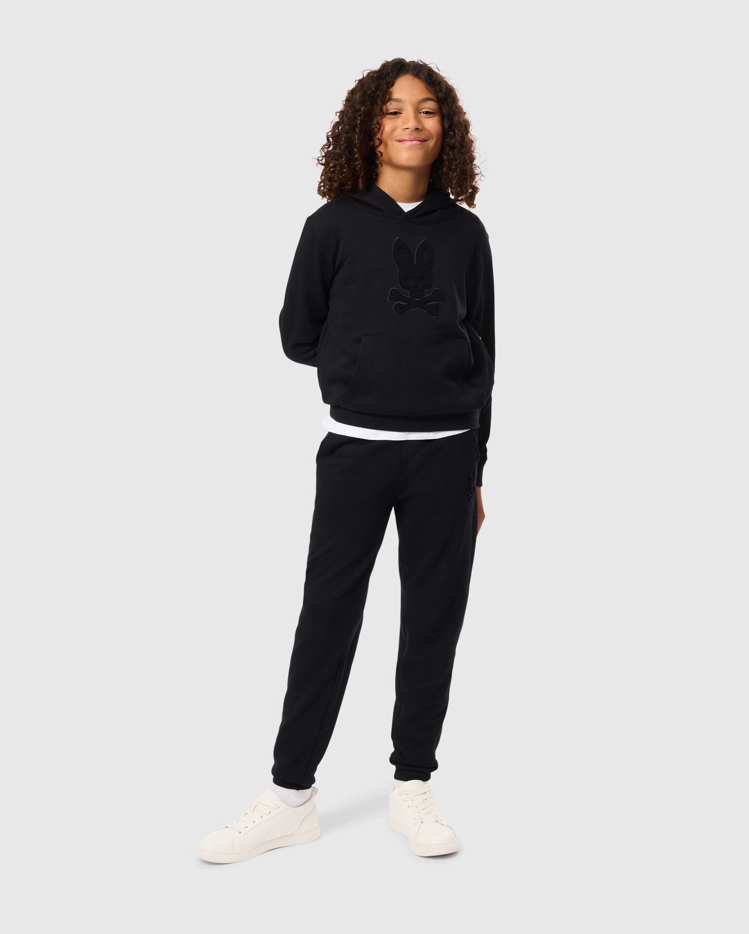 A person with curly hair is standing confidently against a plain background, wearing a black hoodie and the KIDS PACIFIC CHENILLE JOGGER - B0P972D200 by Psycho Bunny. They have their hands behind their back and are smiling, sporting casual bottoms paired with white sneakers.