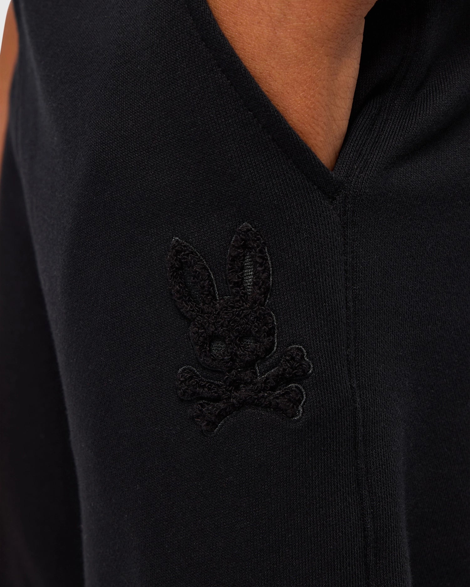 A close-up of a black sleeve features a stitched bunny head above crossbones, with the subtle embroidery seamlessly blending into the fabric, much like the high-pile chenille logo patch on Psycho Bunny's KIDS PACIFIC CHENILLE JOGGER - B0P972D200.