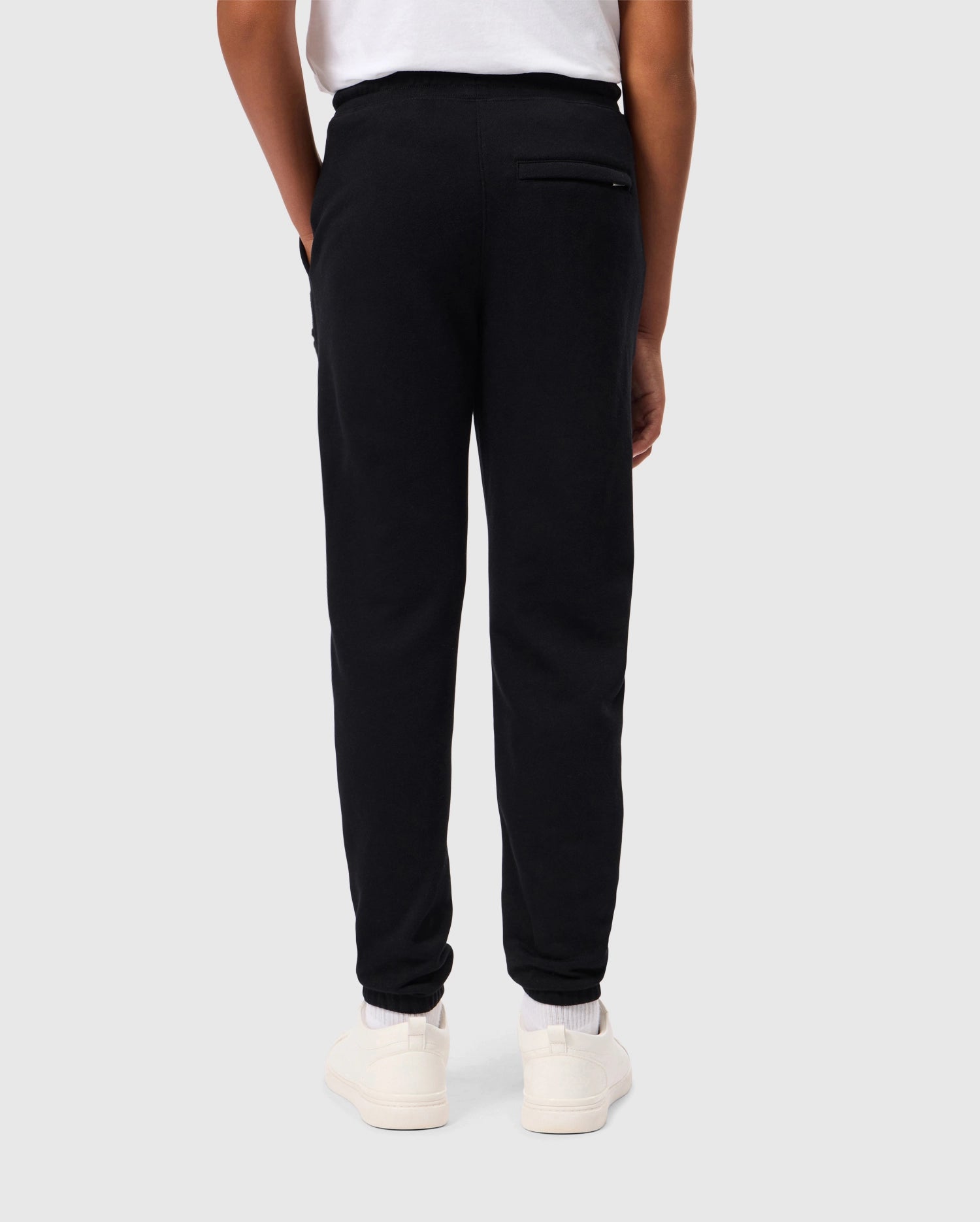 A person dressed in the Psycho Bunny's KIDS PACIFIC CHENILLE JOGGER stands with their back to the camera, wearing a white shirt and matching white sneakers. They casually tuck their hands into their pockets against a plain, light gray background.